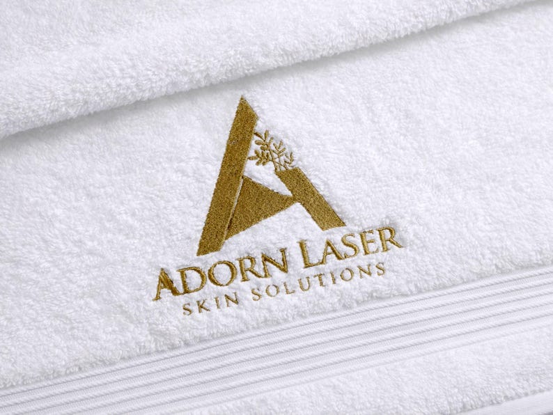 White towel featuring an elegant triangular monogram emblem embroidered in gold thread, a refined laser-cut style logo design.