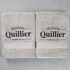 Two beige embroidered towels stacked together, each featuring matching wordmark-style lettering stitched in tonal thread for a coordinated set.