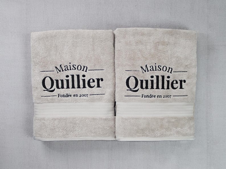 Two beige embroidered towels stacked together, each featuring matching wordmark-style lettering stitched in tonal thread for a coordinated set.