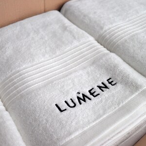 Grey embroidered towel with small lettering stitched along the ribbed border, a subtle minimalist wordmark-style design.