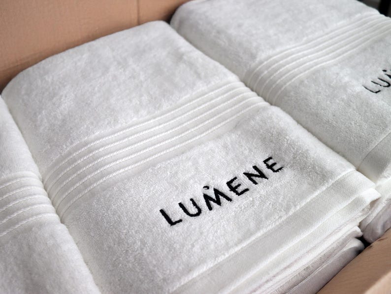 Grey embroidered towel with small lettering stitched along the ribbed border, a subtle minimalist wordmark-style design.