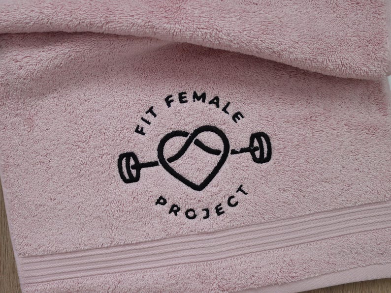 Blush pink embroidered towel featuring a circular fitness badge design with a heart and dumbbell icon, a fun personalised choice for a gym gift.
