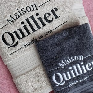 Beige wordmark-embroidered towel styled alongside a navy towel with a woven label detail, shown together to compare embroidery styles and colourways.