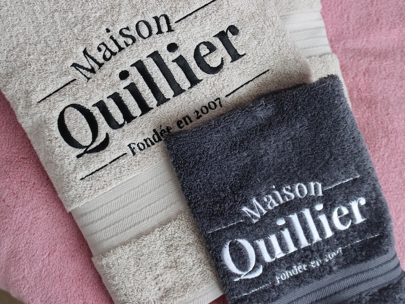 Beige wordmark-embroidered towel styled alongside a navy towel with a woven label detail, shown together to compare embroidery styles and colourways.