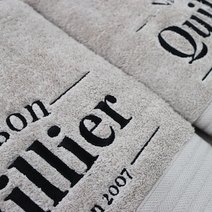 Beige embroidered towel with large bold serif lettering stitched diagonally across the pile, shown close-up to highlight the scale and texture of the embroidery.