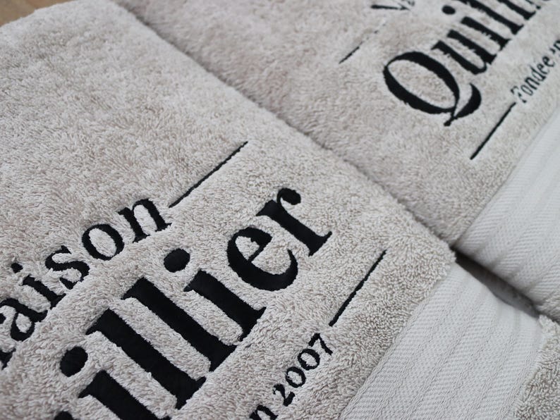 Beige embroidered towel with large bold serif lettering stitched diagonally across the pile, shown close-up to highlight the scale and texture of the embroidery.