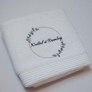 White embroidered towel featuring a circular wreath emblem and script lettering, a minimalist monochrome design folded flat.