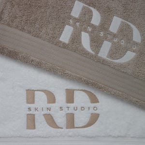 Beige embroidered towel with a bold monogram design stitched in matching thread, folded flat for a clean close-up product shot.