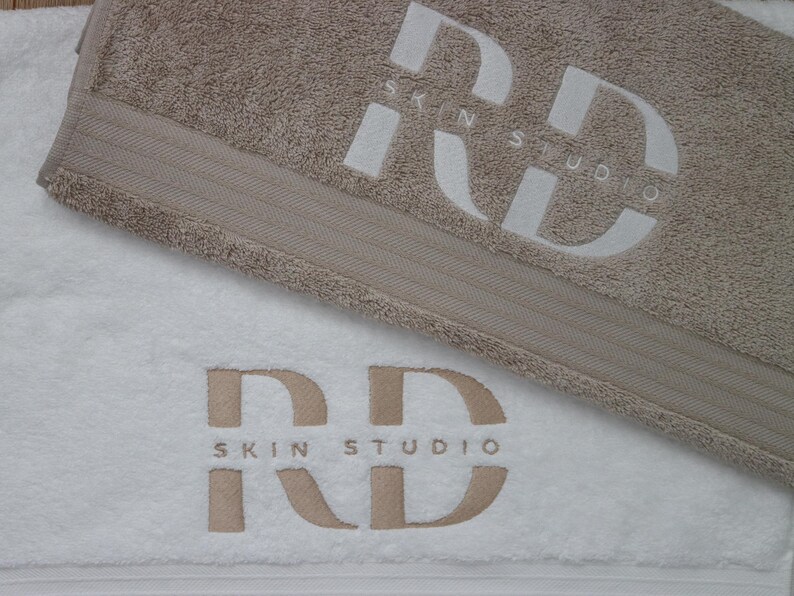 Beige embroidered towel with a bold monogram design stitched in matching thread, folded flat for a clean close-up product shot.