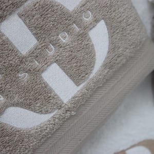 Beige wordmark-embroidered towel styled alongside a navy towel with a woven label detail, shown together to compare embroidery styles and colourways.