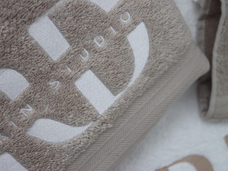 Beige wordmark-embroidered towel styled alongside a navy towel with a woven label detail, shown together to compare embroidery styles and colourways.