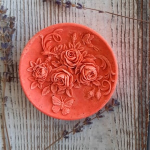 May include: A round, coral-colored dish with a raised design of roses and a butterfly. The dish is made of a smooth, textured material.