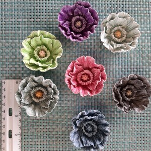 May include: Seven ceramic poppy flower decorations in various colors, including green, purple, pink, gray, and blue. The flowers are arranged on a blue and white patterned surface.