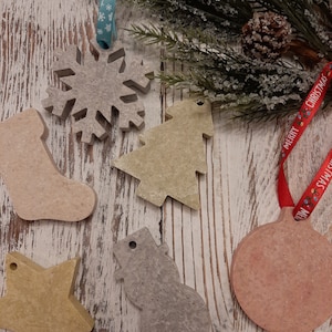 May include: A set of six Christmas ornaments made of concrete. The ornaments are shaped like a snowflake, a stocking, a Christmas tree, a snowman, a star, and a ball. The ornaments are painted in various shades of beige, gray, and pink. The ornaments are displayed on a white wooden background with a sprig of greenery and a red ribbon with the words "Merry Christmas" printed on it.
