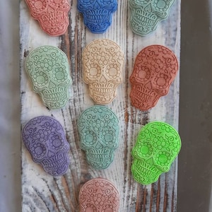 May include: A collection of colorful, decorative skull-shaped objects. The skulls are in various colors, including red, blue, green, and purple, and are adorned with floral patterns. They are arranged on a weathered, white wooden surface.