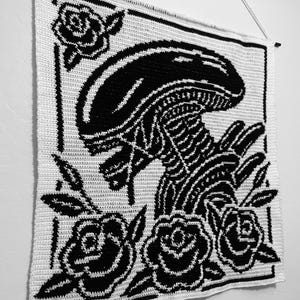 以下が含まれることがあります： 黒と白の編み物ウォールハンギング。エイリアンのデザインとバラが描かれています。作品は黒い縁で縁取られ、白いコードが付いた木の棒から吊り下げられています。ユニークな壁の装飾です。