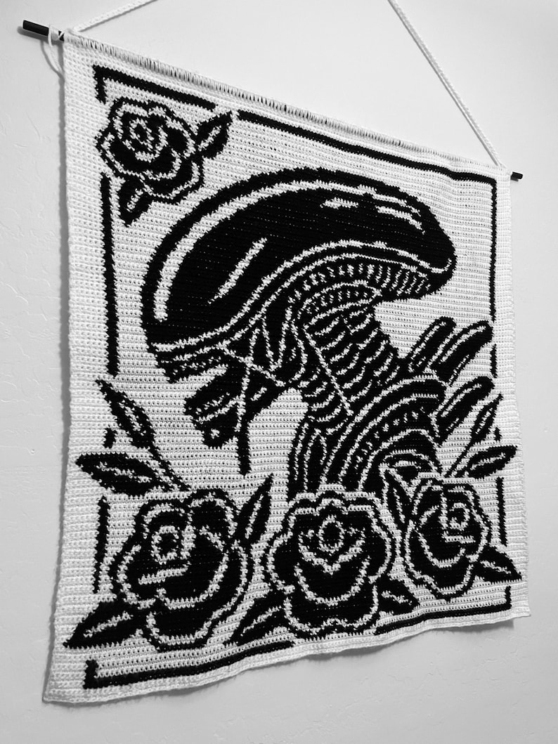 以下が含まれることがあります： 黒と白の編み物ウォールハンギング。エイリアンのデザインとバラが描かれています。作品は黒い縁で縁取られ、白いコードが付いた木の棒から吊り下げられています。ユニークな壁の装飾です。