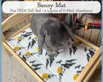May include: A gray bunny rabbit sitting on a white and gray patterned bed with a yellow sun design. The bed is made of wood and has a wooden frame. The text "Bunny Mat" is written in black on a white background above the image. The text "Fits IKEA Doll Bed - 2 Layers of U-Haul Absorbancy" is written in black on a white background below the image.