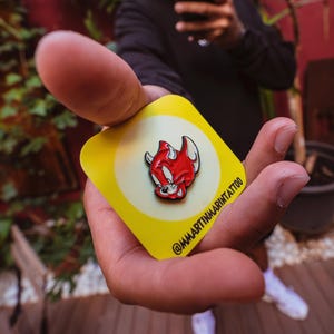 Puede incluir: Un pin de esmalte amarillo y blanco con un diablo de dibujos animados rojo con cuernos y un contorno negro. El pin está sujeto en una mano y el texto "@martinamartintattoo" es visible en la esquina inferior derecha del pin.