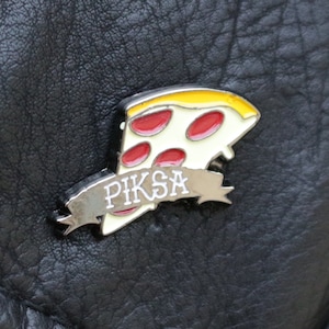 May include: A black leather jacket with a silver button and a metal pin featuring a red and yellow pizza slice with the word "Piksa" on a white banner.
