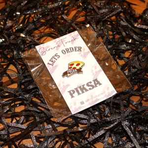 May include: A white card with the text "Siren's Touch Lets Order... Piksa" and a small enamel pin of a pizza slice with the text "Piksa" on it. The pin is in a clear plastic bag.
