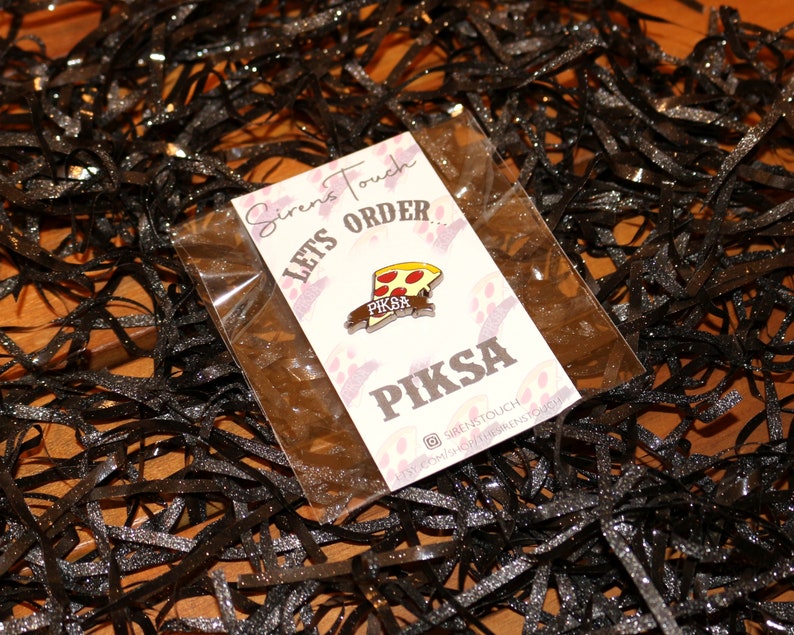 May include: A white card with the text "Siren's Touch Lets Order... Piksa" and a small enamel pin of a pizza slice with the text "Piksa" on it. The pin is in a clear plastic bag.