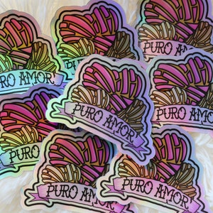 May include: A stack of iridescent heart-shaped stickers with the phrase "Puro Amor" in a banner below each heart. The hearts are filled with a colorful, geometric pattern.