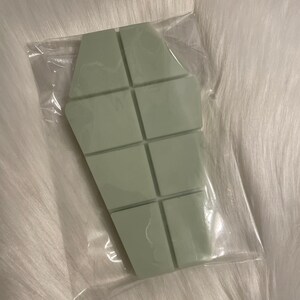 May include: A light green wax melt in the shape of a coffin, divided into 10 squares. The wax melt is packaged in a clear plastic bag.