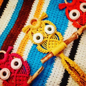 May include: Three colourful macrame owl wall hangings with wooden dowels. The owls are red, yellow, and pink. The owls are hanging on a white and blue striped crocheted background.