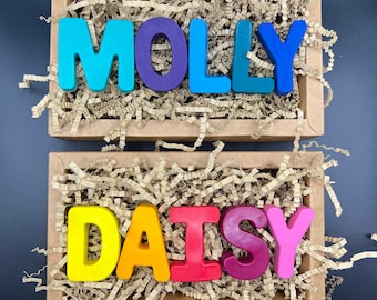 Puede incluir: Dos cajas llenas de papel picado, cada una con un nombre escrito en letras de cera de colores. La caja superior contiene el nombre "MOLLY" en letras azules, moradas, azules, verdes y azules. La caja inferior contiene el nombre "DAISY" en letras amarillas, naranjas, rojas, burdeos y rosas.