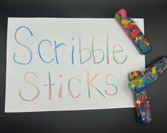 May include: A white paper with the words "Scribble Sticks" written in colorful crayon. Three multi-colored crayons are positioned around the paper. The crayons are a mix of colors, including blue, red, yellow, and green.