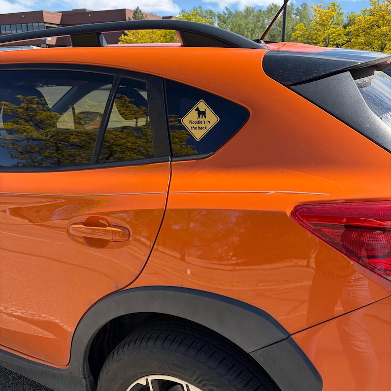 May include: An orange car with black trim and a window sticker. The yellow sticker has a black dog silhouette and the text "Noodle's in the back".