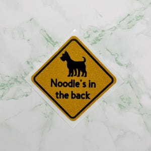May include: A diamond-shaped sticker with a gold glitter background and a black border. The sticker features a black silhouette of a dog and the text "Noodle's in the back" in black font. The sticker is on a white background.