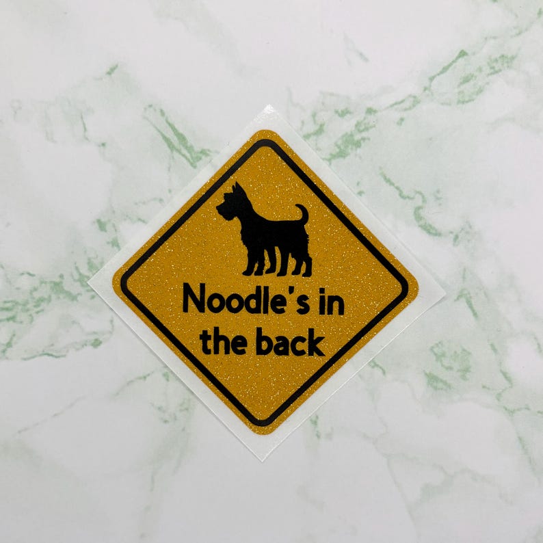 May include: A diamond-shaped sticker with a gold glitter background and a black border. The sticker features a black silhouette of a dog and the text "Noodle's in the back" in black font. The sticker is on a white background.