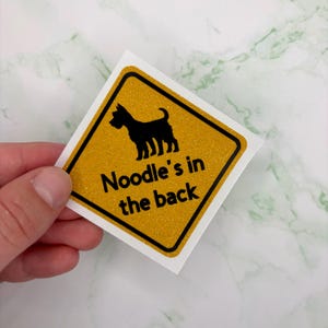 May include: A square sticker with a yellow glitter background and a black border. The sticker features a black silhouette of a dog and the text "Noodle's in the back" in black.