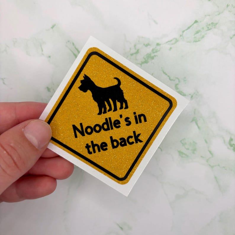 May include: A square sticker with a yellow glitter background and a black border. The sticker features a black silhouette of a dog and the text "Noodle's in the back" in black.