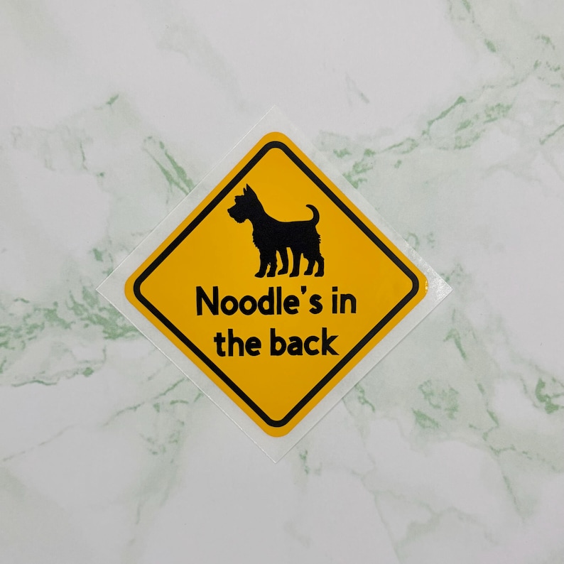 May include: A yellow diamond-shaped sticker with a black border and a black dog silhouette. The text "Noodle's in the back" is printed in black below the dog illustration. The sticker is on a white and green marble surface.