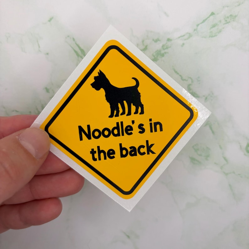 May include: A yellow diamond-shaped sticker featuring a black dog silhouette and the text "Noodle's in the back". The sticker has a black border. The sticker is held by a hand.