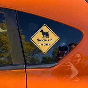 May include: A yellow diamond car sticker with a black dog silhouette and the text "Noodle's in the back". The sticker is on a car window, with an orange car body visible.