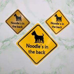 May include: Three yellow diamond stickers with black borders, each featuring a black dog silhouette and the text "Noodle's in the back". One sticker has a gold glitter background.
