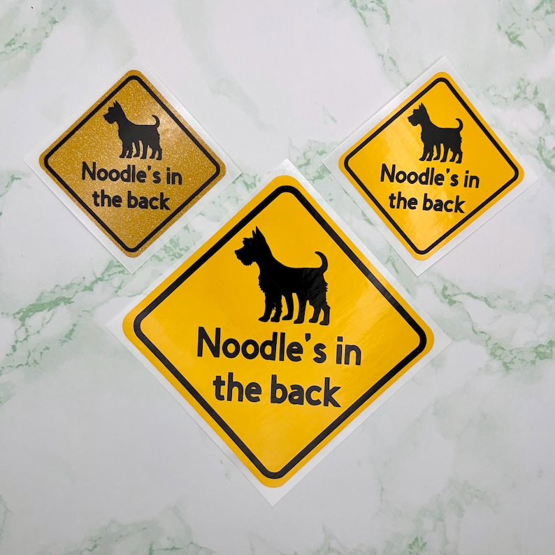 May include: Three yellow diamond stickers with black borders, each featuring a black dog silhouette and the text "Noodle's in the back". One sticker has a gold glitter background.