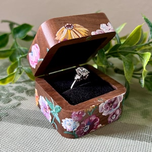 May include: A wooden ring box with a hinged lid, open to reveal a diamond engagement ring. The box is painted with colorful floral designs in pink, purple, yellow, and white. The interior is lined with black velvet.