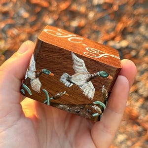 May include: A small, rectangular wooden box featuring a hand-painted scene of two mallard ducks in flight over cattails. The top of the box displays the white cursive lettering H.F. The wood has a rich brown grain, and the painted details include green, white, and brown tones.