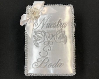 May include: White satin wedding ring pillow with silver embroidery. The pillow features the words "Nuestra Boda" and a design of two doves. A white bow with a rhinestone embellishment is attached to the top.