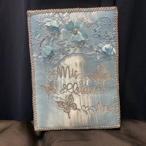 May include: A light blue satin keepsake box with silver embroidery. The box features a lace design with flowers and butterflies. The text "Mis Quince Años" is embroidered on the front.