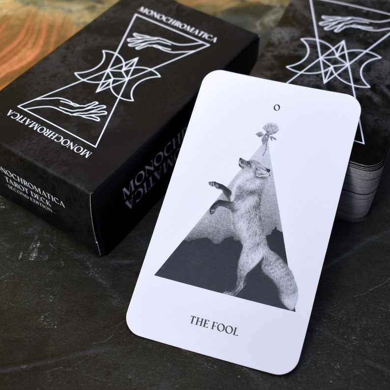 May include: A black and white tarot card with a fox standing on a triangle. The card is titled "The Fool" and has a rose with a stem at the top. The card is from the Monochromatica Tarot Deck.