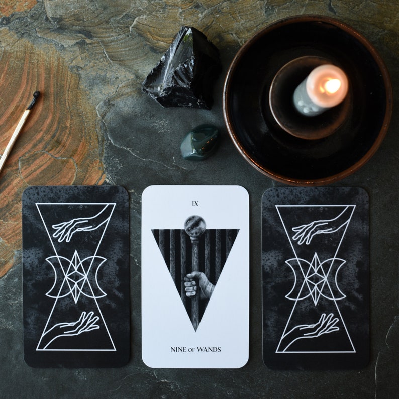 May include: Three tarot cards with black and white illustrations. The middle card is white with black illustrations and reads "IX NINE OF WANDS". The cards are on a dark gray surface with a black stone and a green stone.