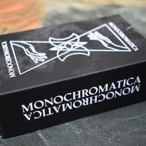 May include: A black box with the word "MONOCHROMATICA" printed in white on the front and sides. The box features a graphic design of two hands reaching towards each other, a star, and two crescent moons.