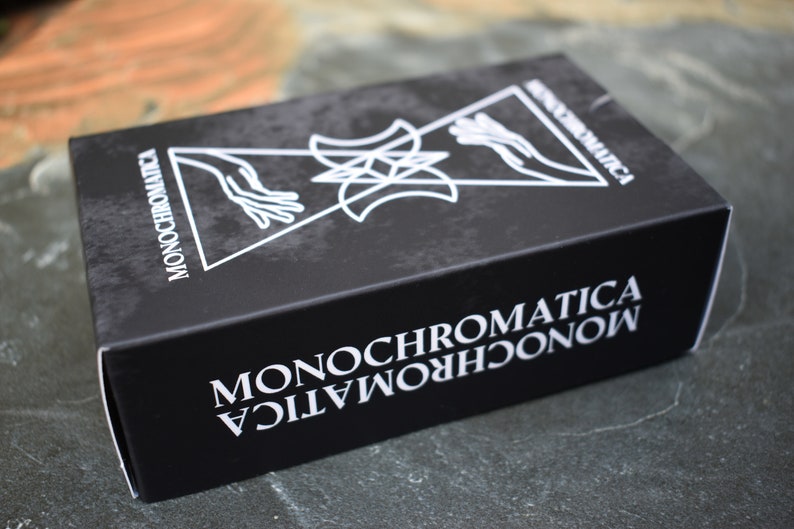 May include: A black box with the word "MONOCHROMATICA" printed in white on the front and sides. The box features a graphic design of two hands reaching towards each other, a star, and two crescent moons.
