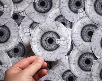 May include: A close-up of a hand holding a white sticker with a black eye design. The sticker has a textured surface and looks like a human eye with veins.